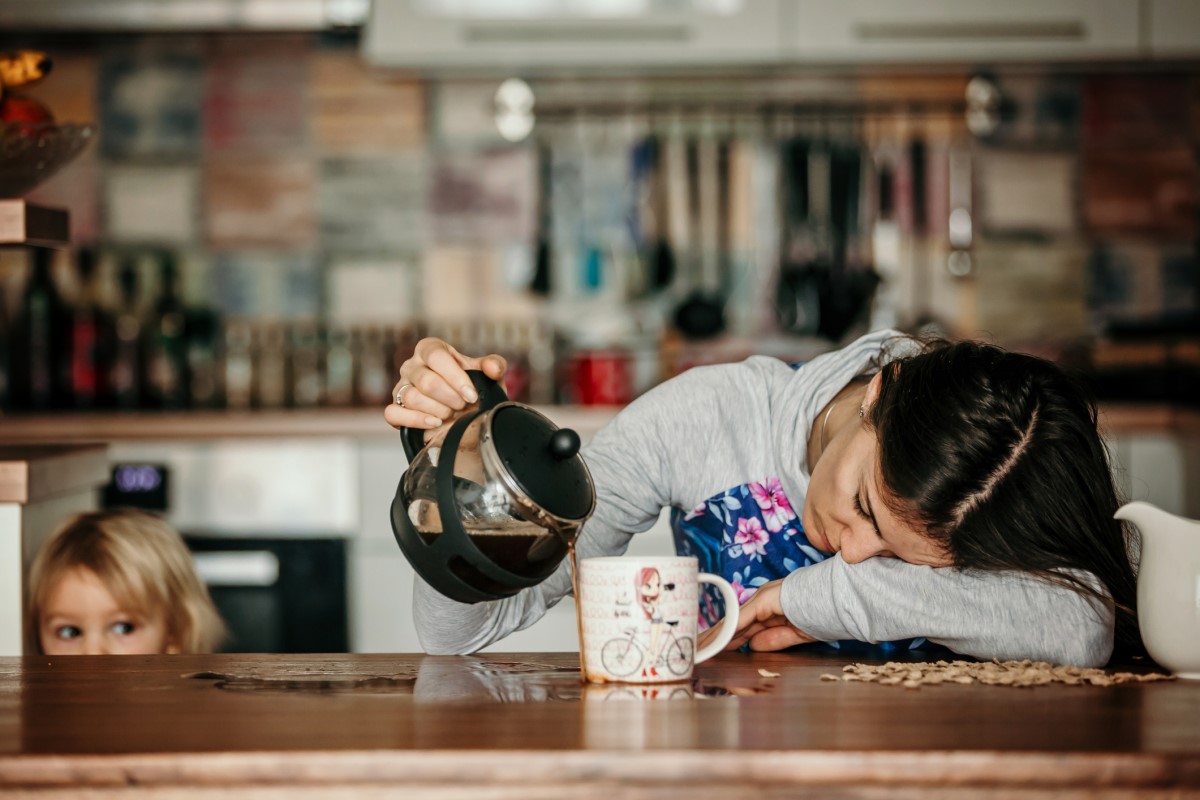 En trött kvinna ligger på köksbänken och spiller kaffe bredvid en mugg, ett barn står bredvid henne.