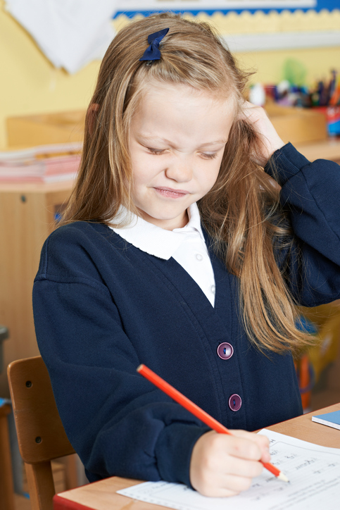 En liten flicka som sitter i skolan med kliande huvud, förmodligen löss i håret, blont hår, sitter och skriver